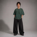 A model stands against a neutral gray studio background wearing an oversized dark green short-sleeve top and very wide-leg black pants.
