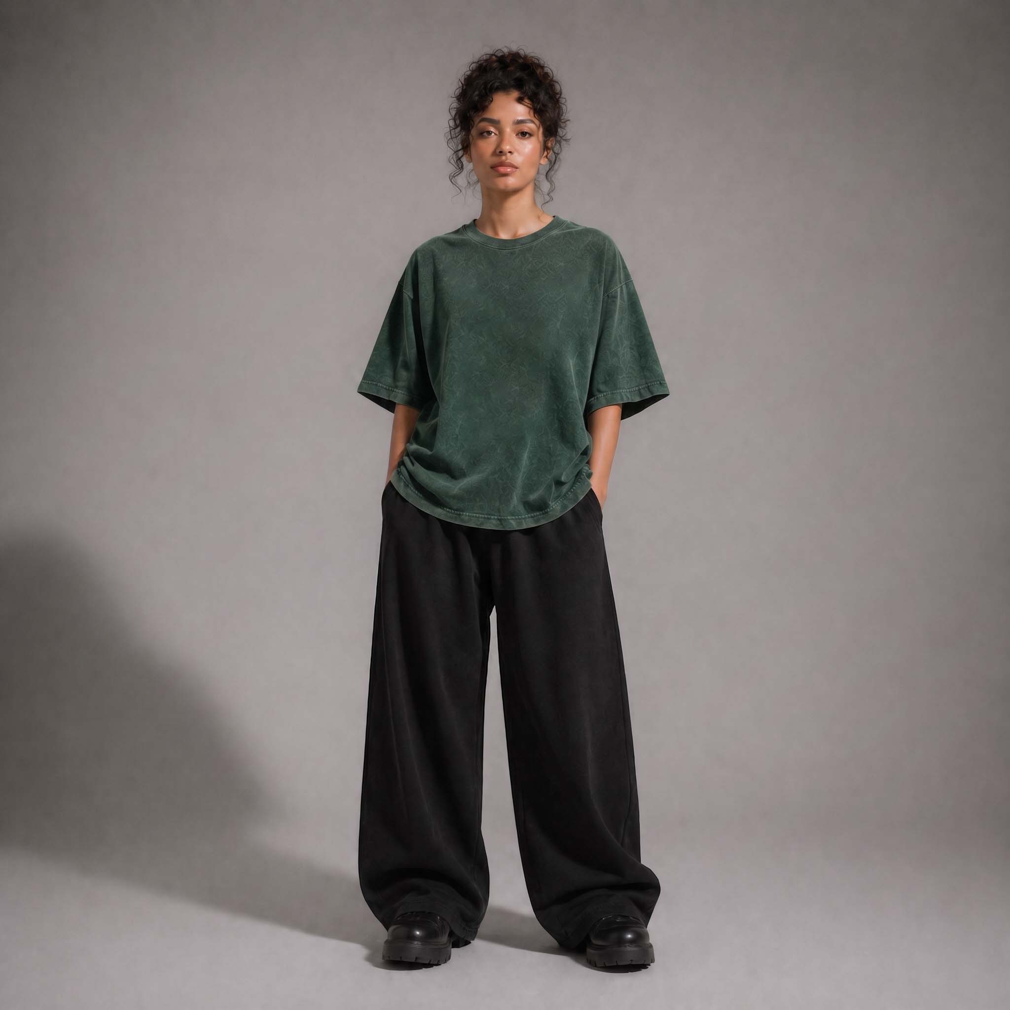 A model stands against a neutral gray studio background wearing an oversized dark green short-sleeve top and very wide-leg black pants.