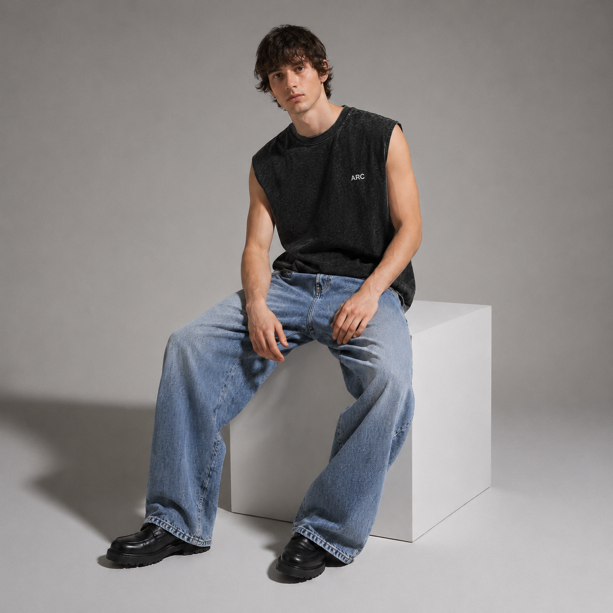 A man sits on a white cube wearing a black sleeveless t-shirt with a small white logo on the chest, light blue wide-leg jeans, and black shoes, against a gray studio background.