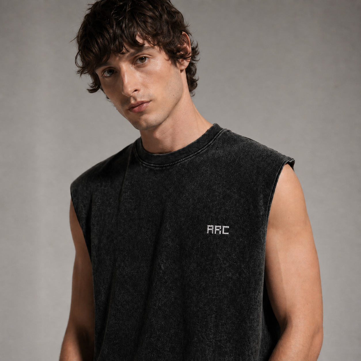 A man wearing a dark charcoal gray sleeveless crewneck t-shirt with a small white 'ARC' logo on the left chest, photographed against a neutral gray background.