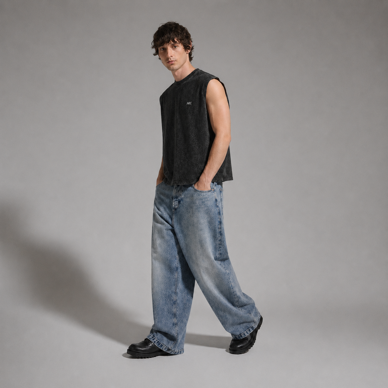 A man wearing a dark charcoal sleeveless t-shirt, loose light-wash blue jeans, and black shoes, standing against a neutral gray studio background.