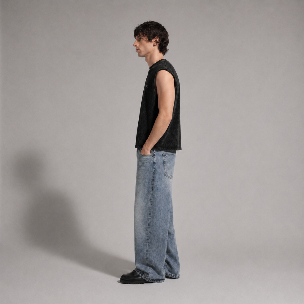 A man in profile wearing a black sleeveless top and light-wash wide-leg jeans, standing against a plain light gray studio background.