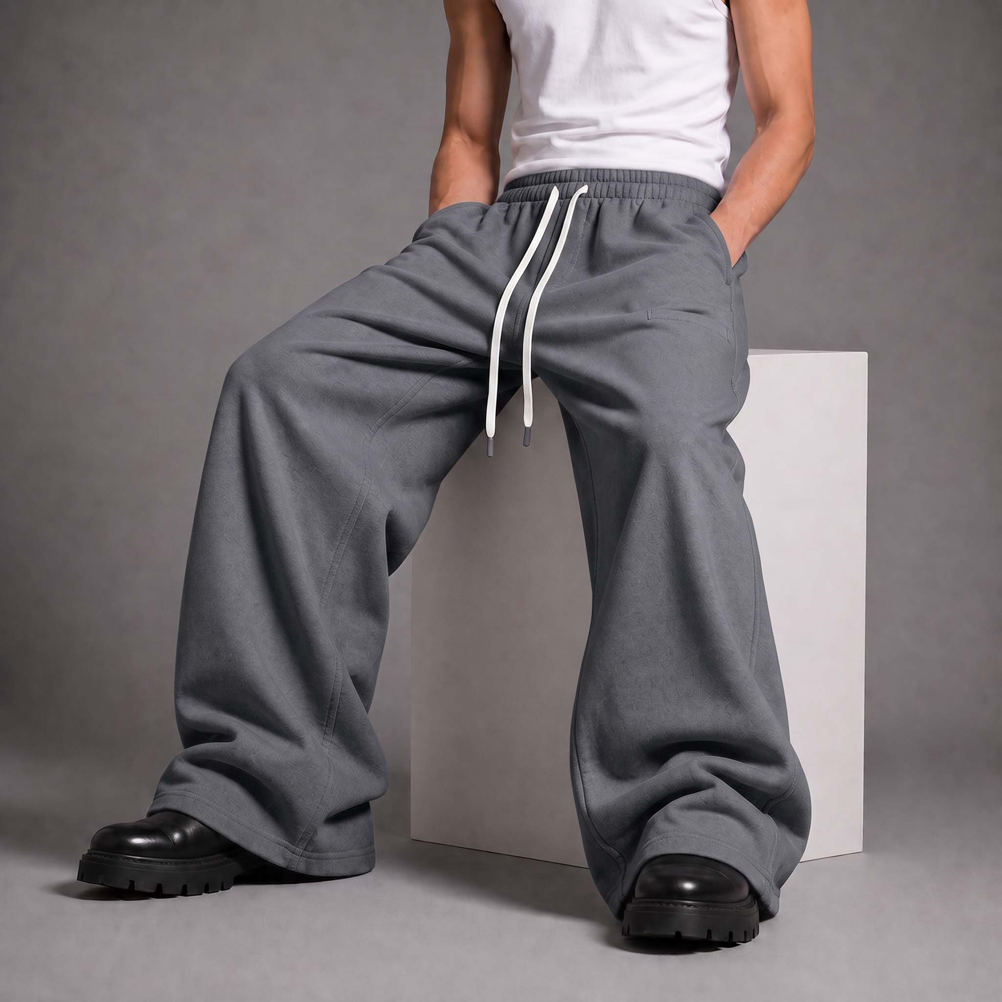A person wearing loose, wide-leg gray sweatpants with a white drawstring, paired with a white tank top and black boots, seated on a white block against a gray studio background.