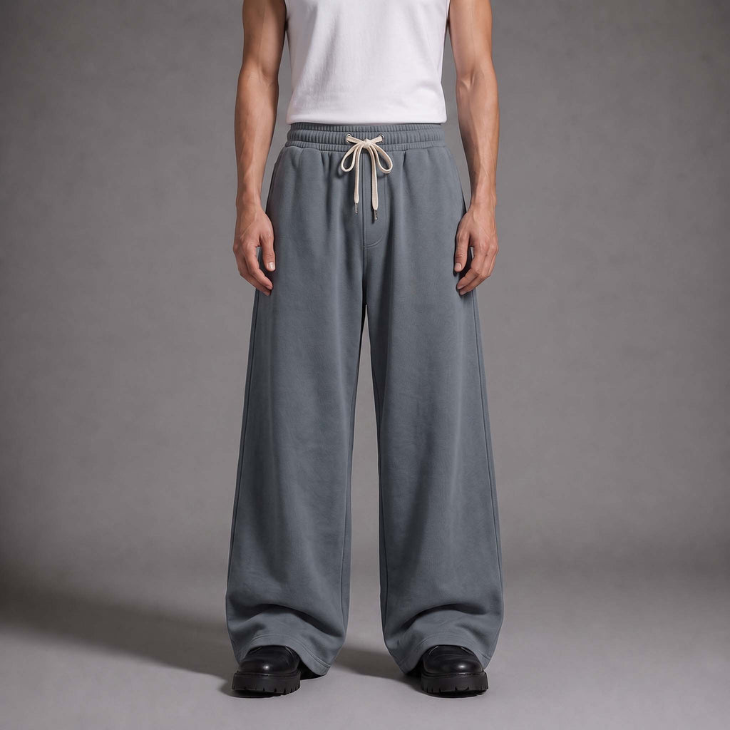 A model from the waist down wearing wide-leg, relaxed-fit gray sweatpants with a cream drawstring, paired with a white tank top and black shoes, against a neutral gray studio background.