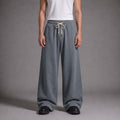 A model from the waist down wearing wide-leg, relaxed-fit gray sweatpants with a cream drawstring, paired with a white tank top and black shoes, against a neutral gray studio background.