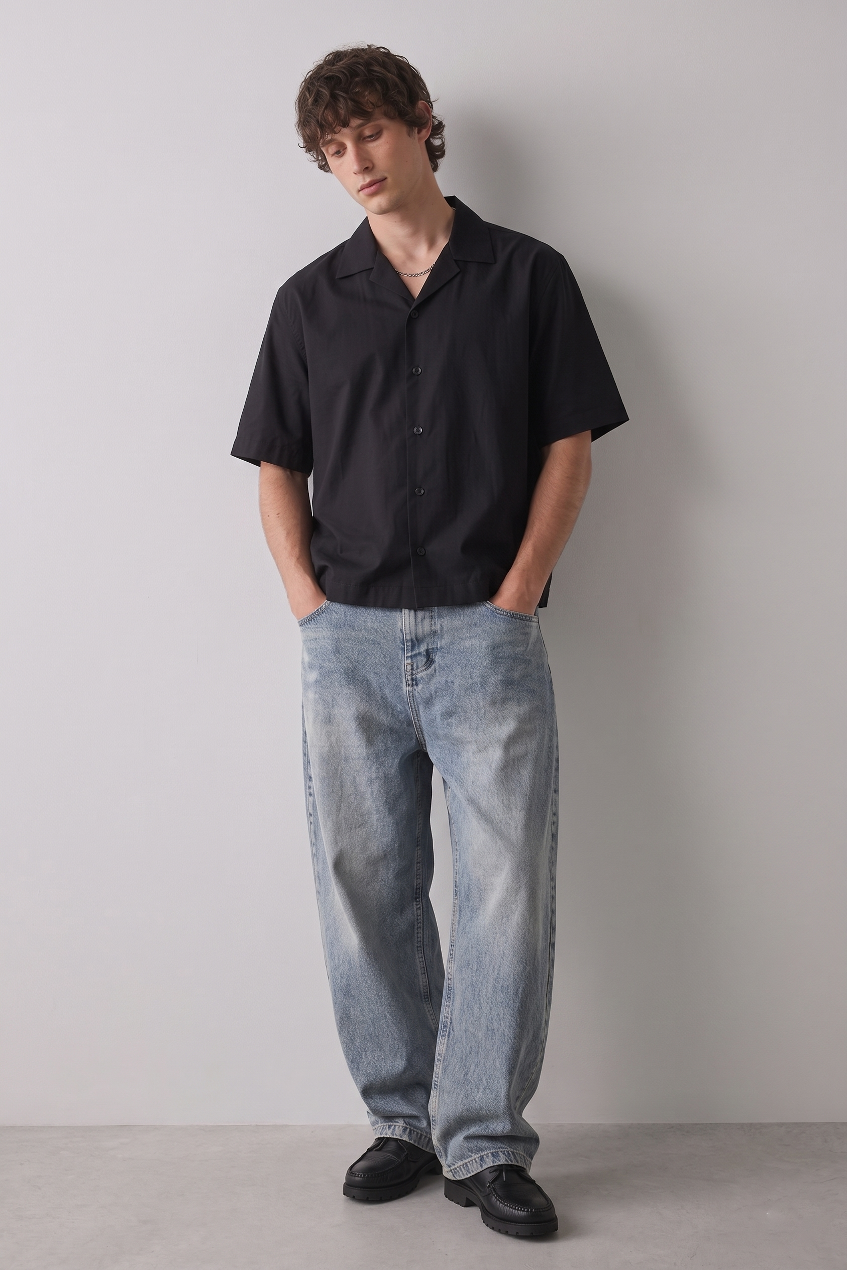 Person wearing a black shirt and light blue jeans against a plain background
Model sitting on a white cube wearing the ARC Black Boxy Fit Shirt in a minimalist lifestyle and fashion editorial setting.