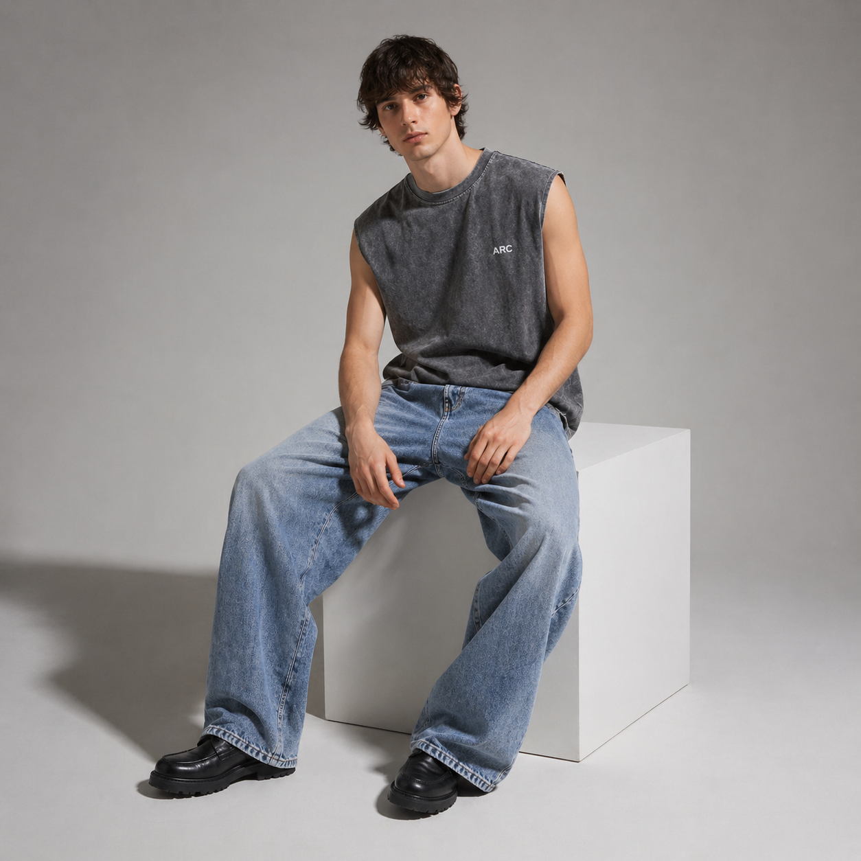 Man wearing a sleeveless gray top and blue jeans sitting on a white cube against a plain background
Model sitting on a white cube wearing the ARC Sleeveless Acid Wash T-Shirt in a casual streetwear lifestyle pose.